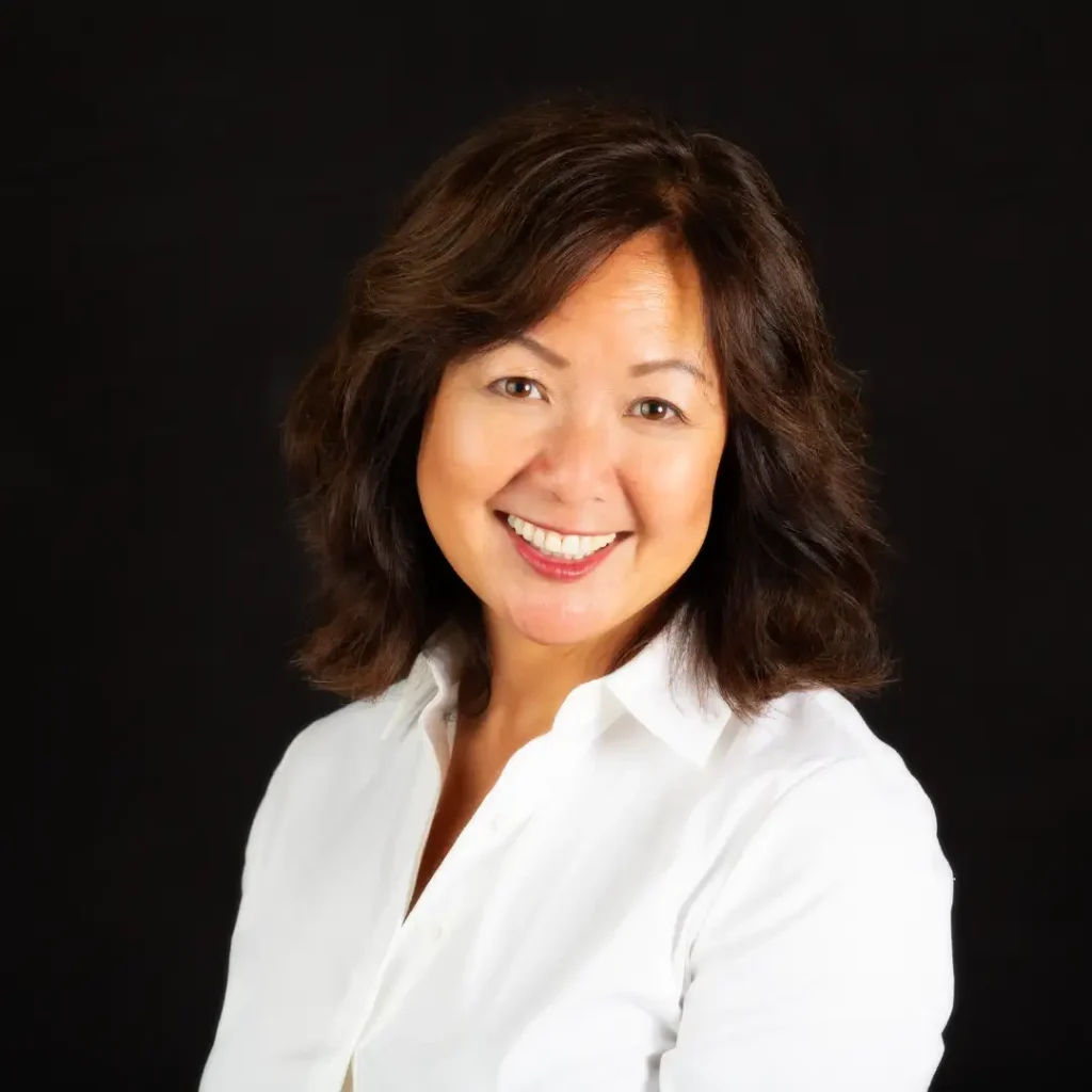 Portrait of a smiling Asian woman with shoulder-length dark hair, wearing a white blouse against a black background, studio headshot.