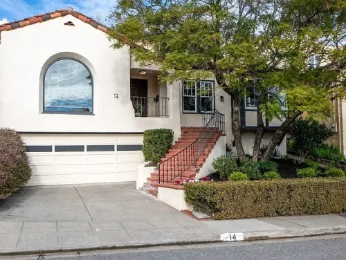 White stucco house with an arched window, brick front steps, and trimmed shrubs by a two-car garage.
