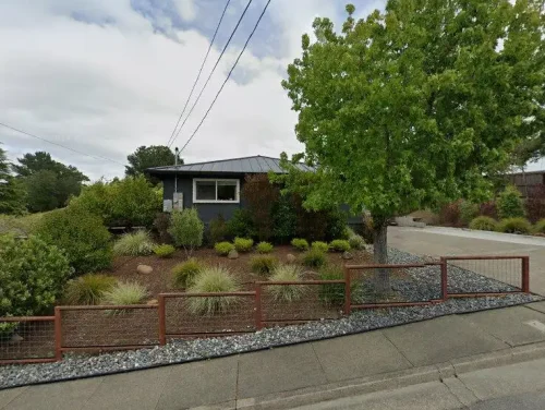 A single-story blue-gray house with a manicured desert-style front yard, red metal fence, and a large tree by the sidewalk.