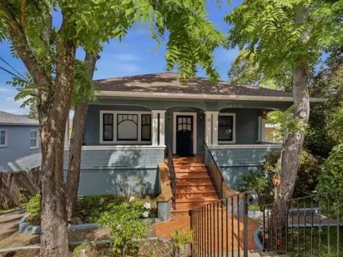 Front view of a blue bungalow with a wooden staircase and black front door, shaded by trees on a sunny day.