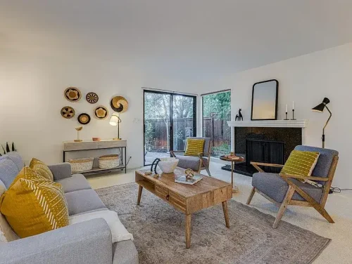 Living room with gray sofa and two armchairs around a wooden coffee table, fireplace with a mirror, and large glass doors to a backyard garden; yellow cushions add color.